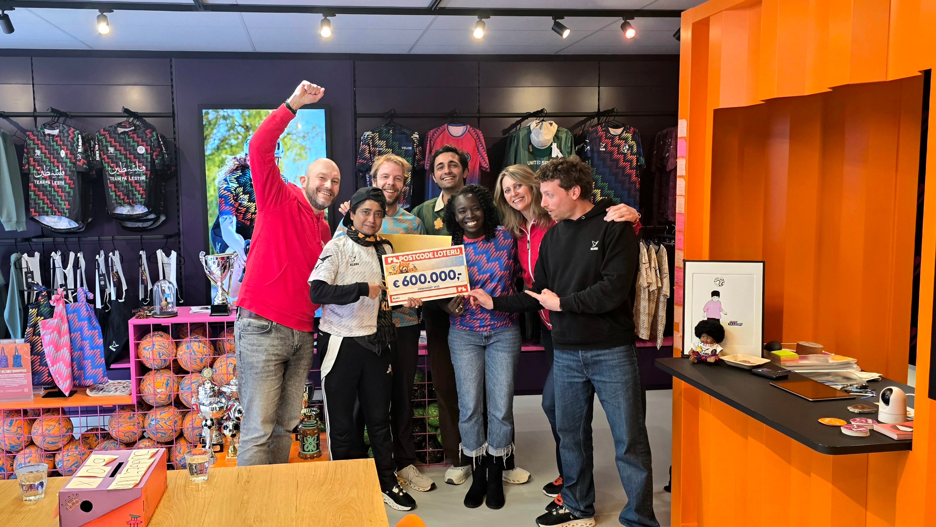 A group of people smiling and holding a large check for €600,000 inside a vibrant, colorful clothing store.